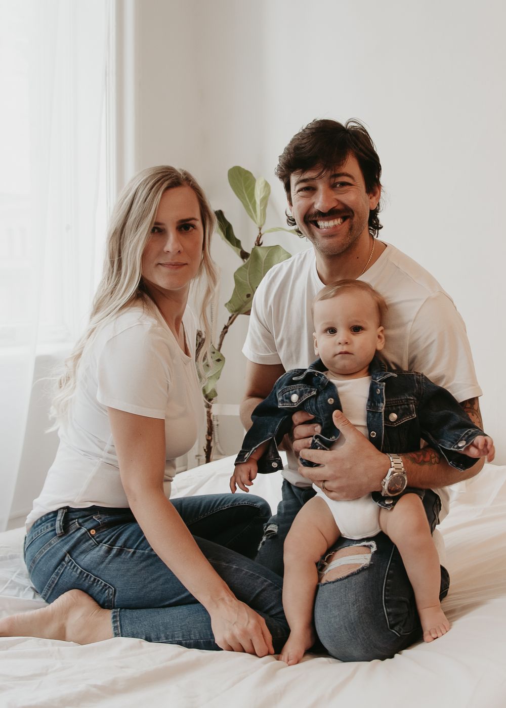 A casual family portrait on a white bed with houseplant in the background.