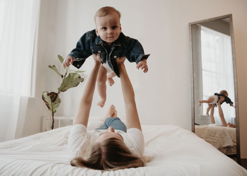 A parent lifting a baby in the air while lying on a white bed.