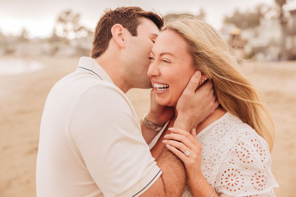 A couple shares a joyful romantic moment on a beach, the man kissing the laughing woman's cheek tenderly.