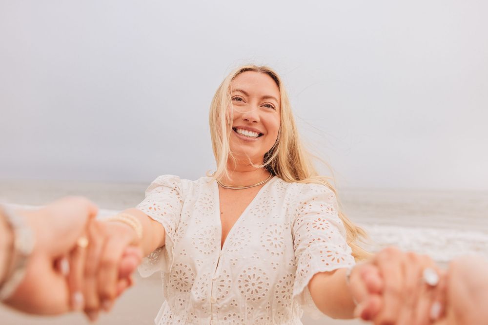 Smiling blonde woman in white lace dress holding hands on a beach, radiating joy and happiness by the ocean.