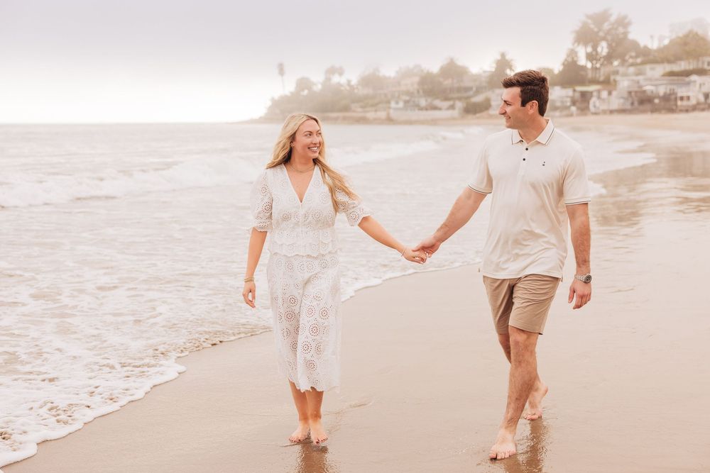 A couple walks hand-in-hand along a sandy beach shore, smiling at each other in casual white and beige outfits.