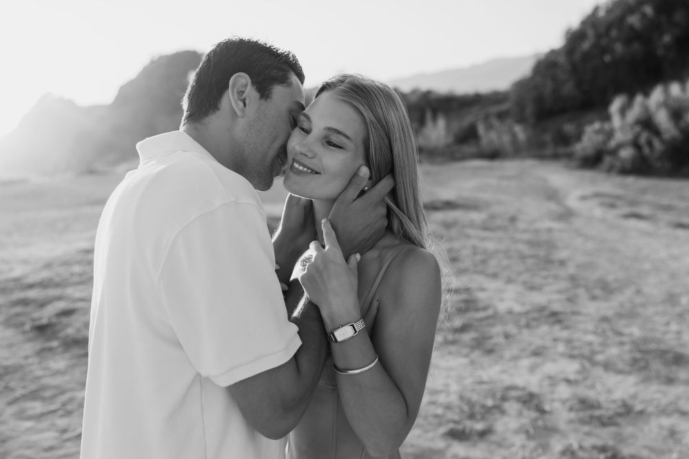 Black and white photo of a couple sharing a tender moment outdoors, man whispering to smiling woman at golden hour.