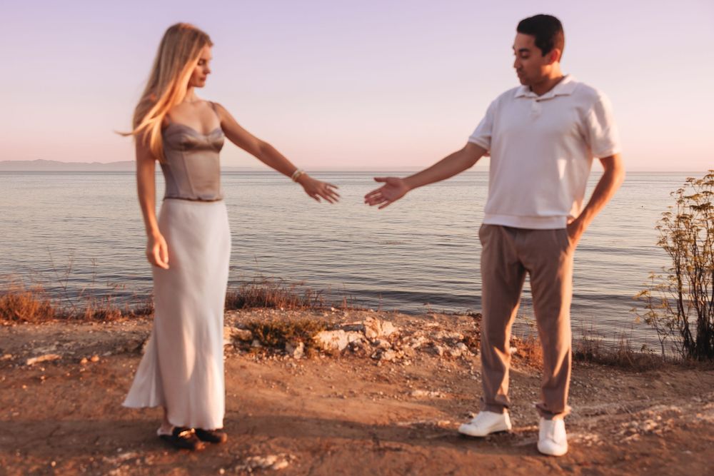 A man and woman reach toward each other near a scenic lakeside at sunset, standing on rocky ground.