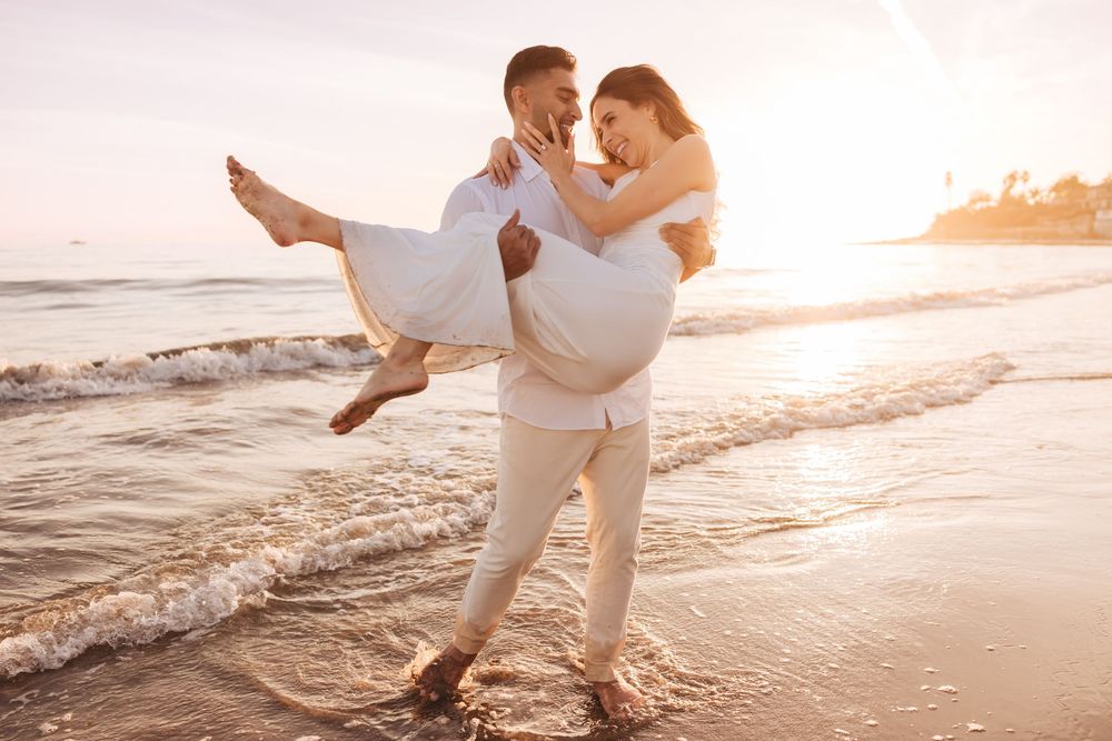 Romantic couple in white outfits embracing on a sunny beach at sunset, man carrying woman in his arms by the ocean waves.