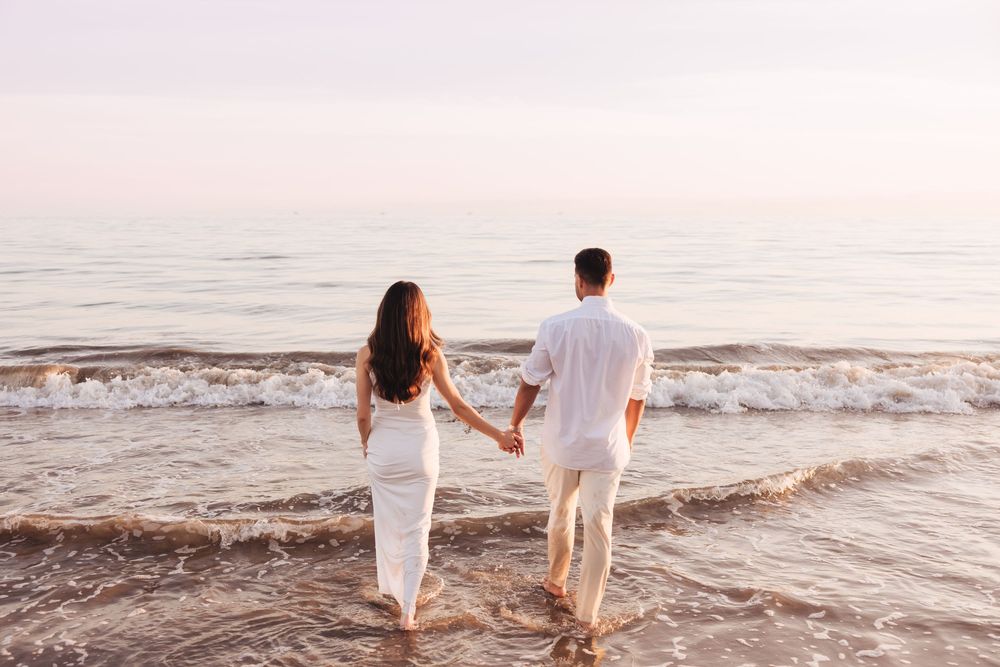 Couple holding hands walking along a serene beach shore at sunset, dressed in white outfits.