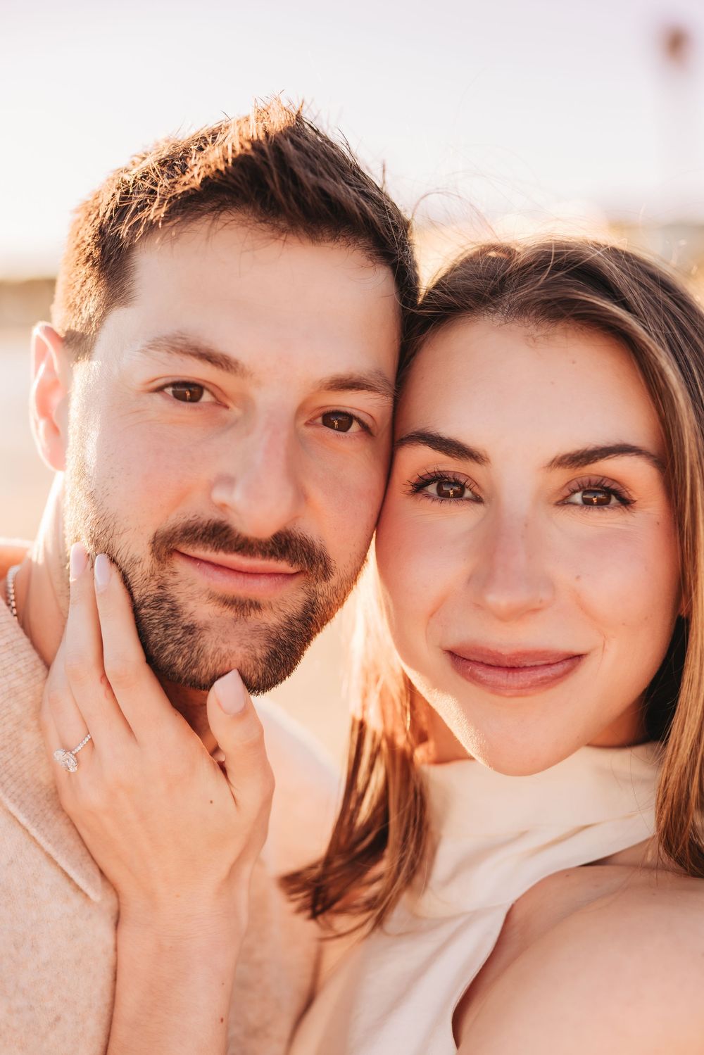 A smiling couple poses cheek to cheek in warm golden light, the woman wearing an engagement ring.