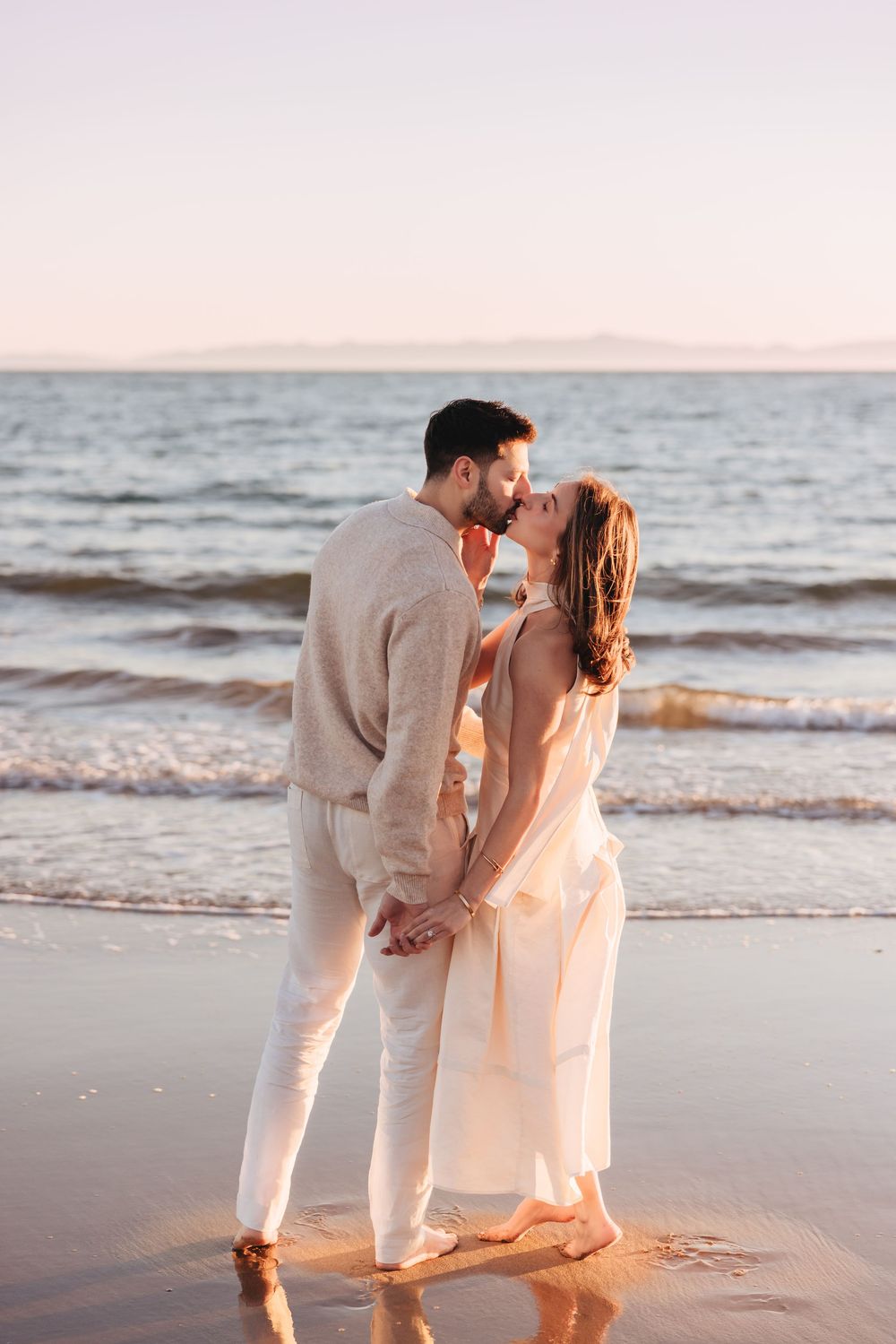 Romantic couple holding hands and kissing at beach sunset, dressed in elegant cream attire by the ocean.