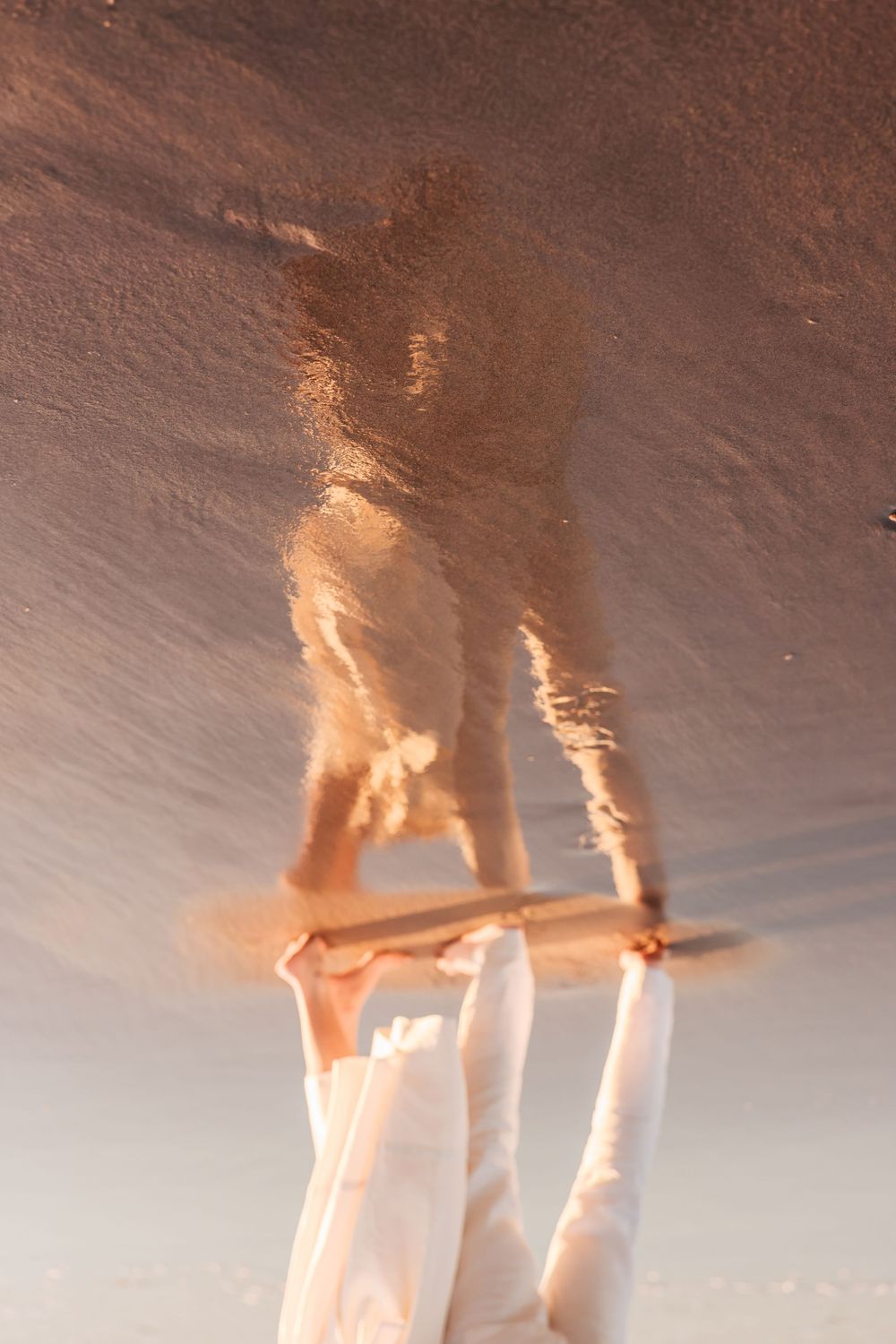 Artistic reflection of a couple holding hands captured in wet beach sand during golden hour sunset.