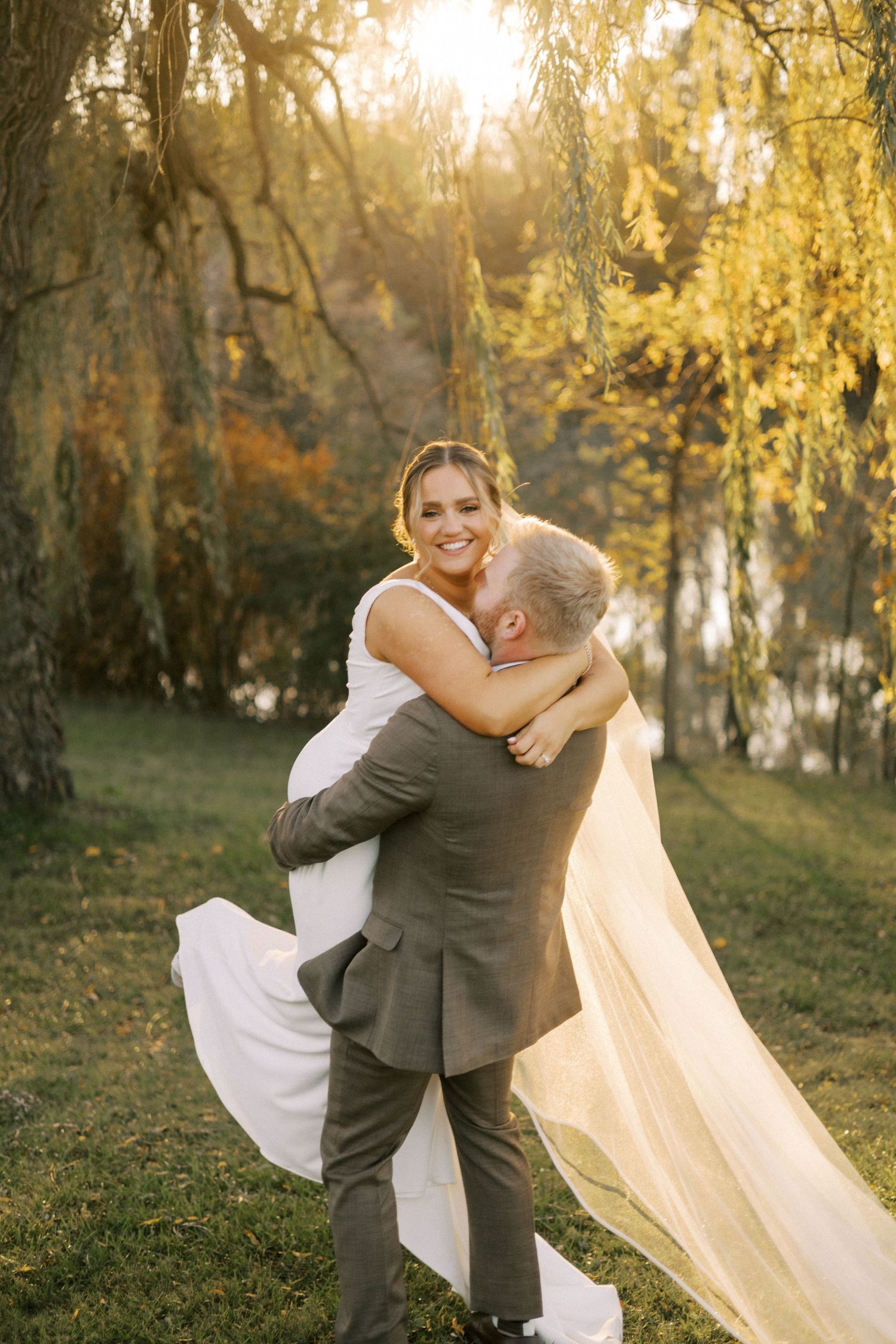 Bride and groom sharing romantic embrace outdoors with sunlit willow trees in background.