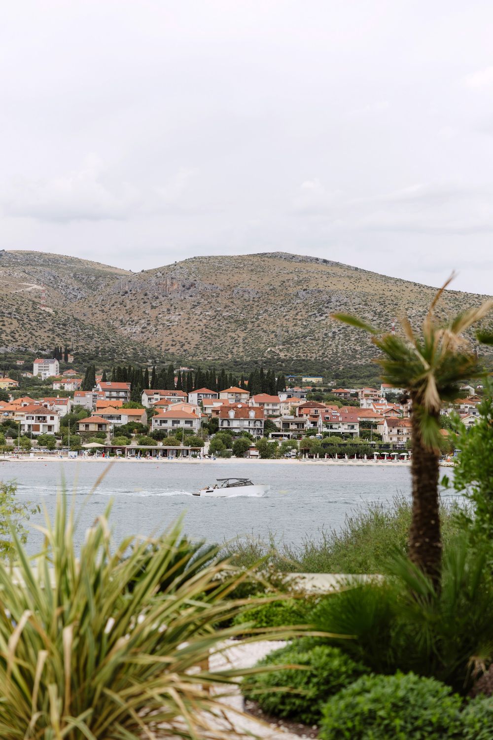 Coastal Mediterranean town with red-roofed buildings nestled along the bay, viewed through tropical palm fronds.