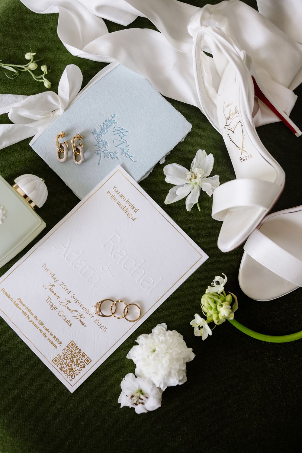 Wedding details flatlay with white invitation, rings, shoes, and flowers on dark green background.