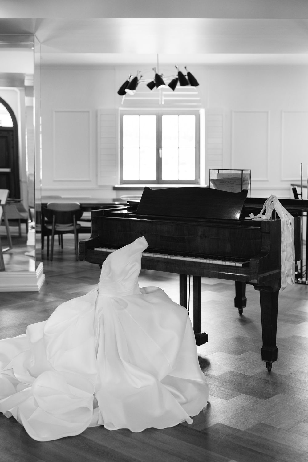 A white wedding dress elegantly draped over a grand piano in a bright room with hardwood floors and pendant lights.