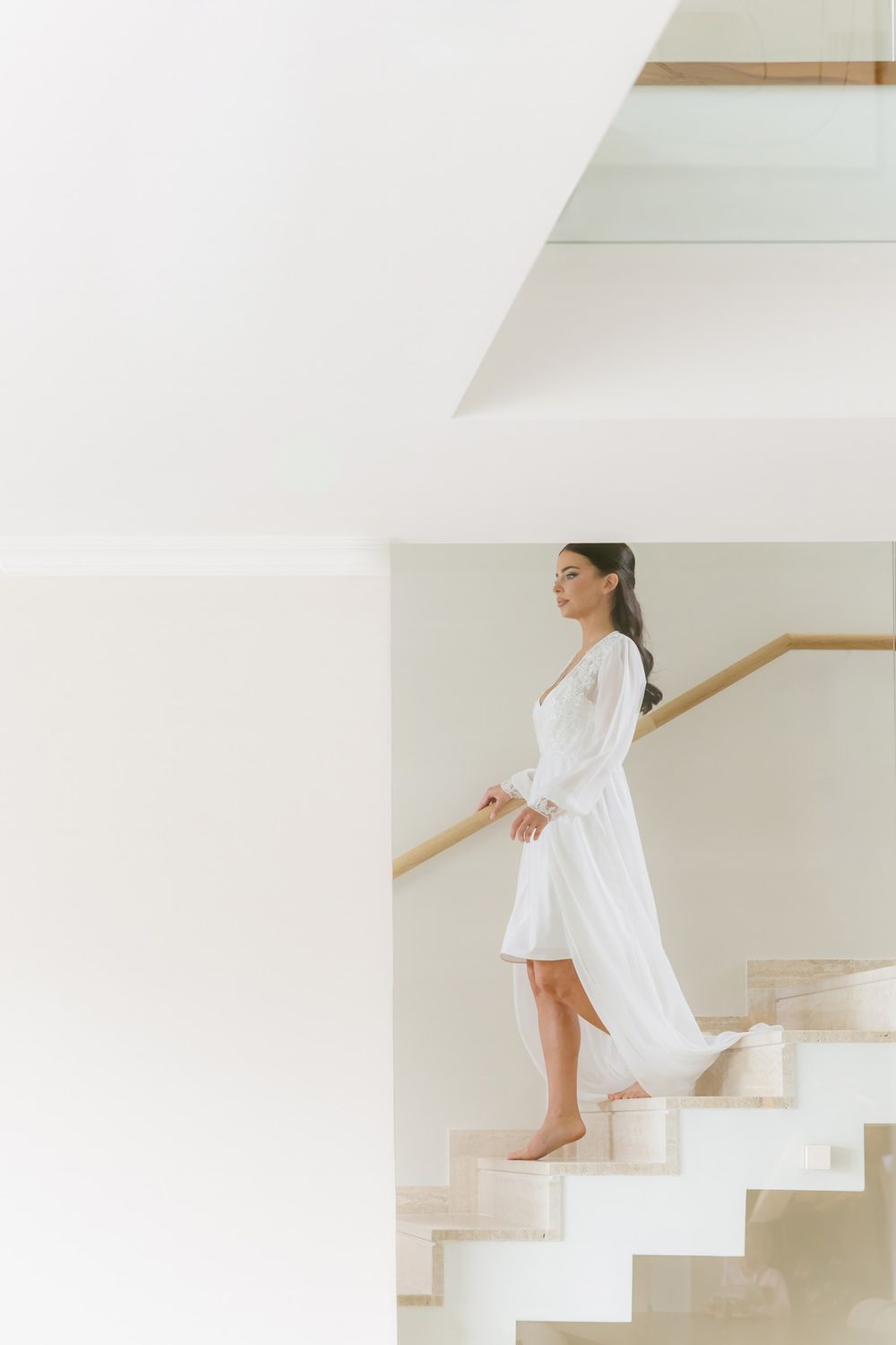 A person in a white outfit sits elegantly on minimalist white stairs with a wooden handrail in a bright interior space.