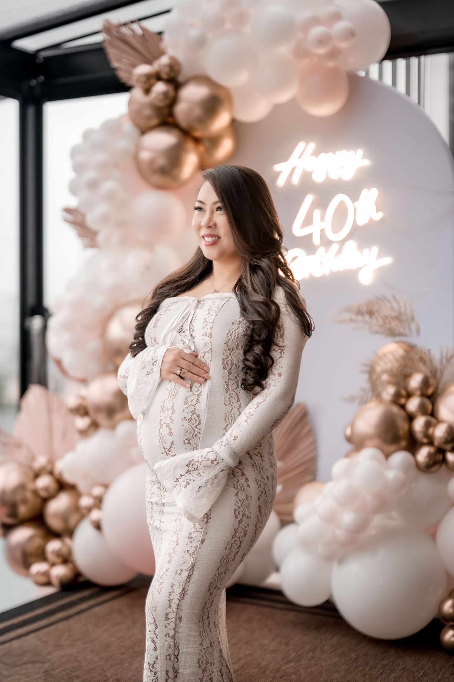 A woman in a white lace dress poses against a backdrop of pink and rose gold balloon decorations at a celebration.