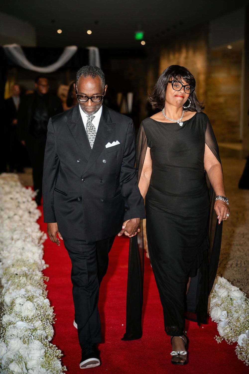 Couples walking down a red carpet lined with white floral arrangements.