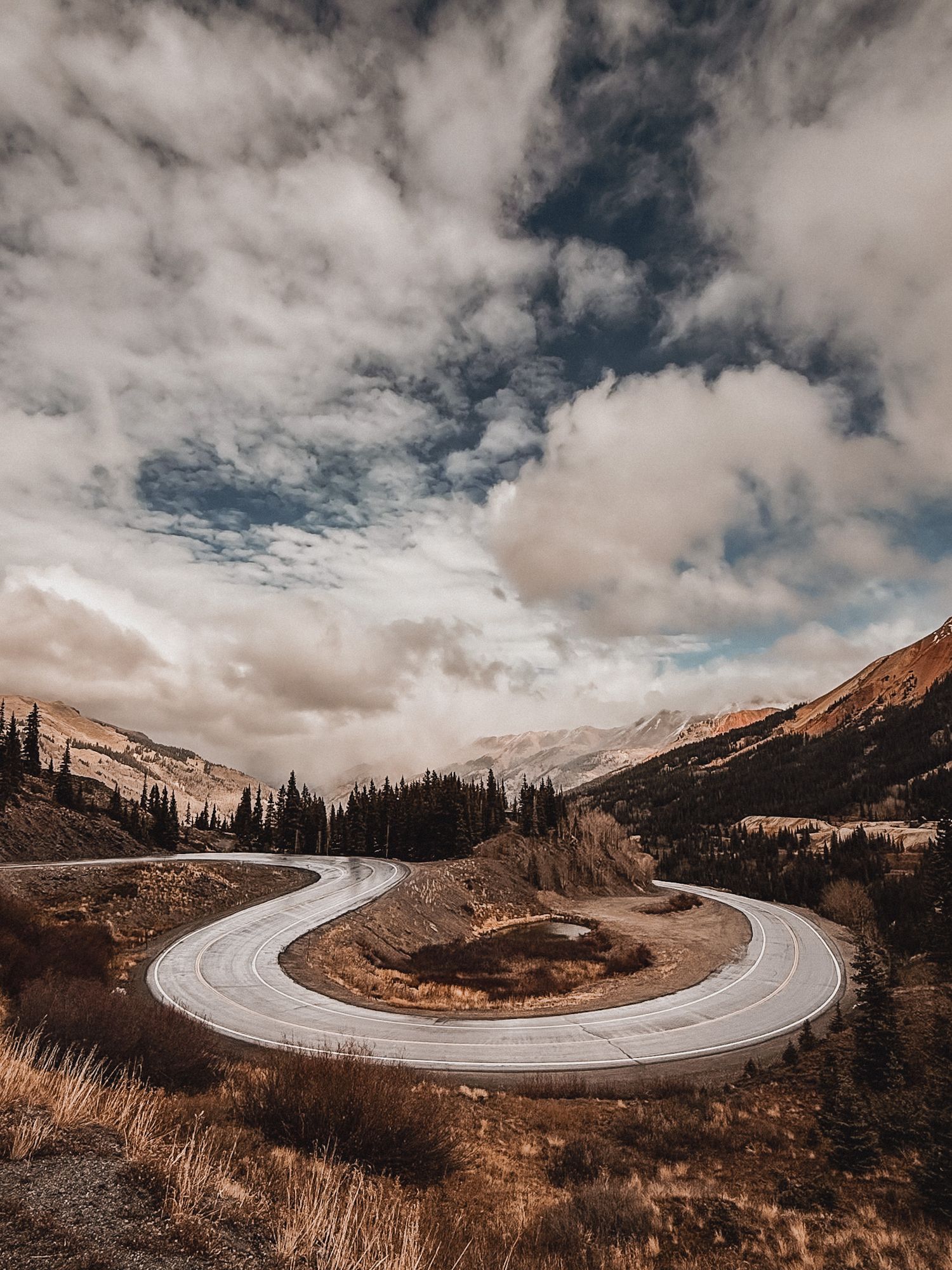 A mountain road in Colorado curves through dramatic mountain landscape under moody cloudy skies in autumn colors. Million Dollar Highway.