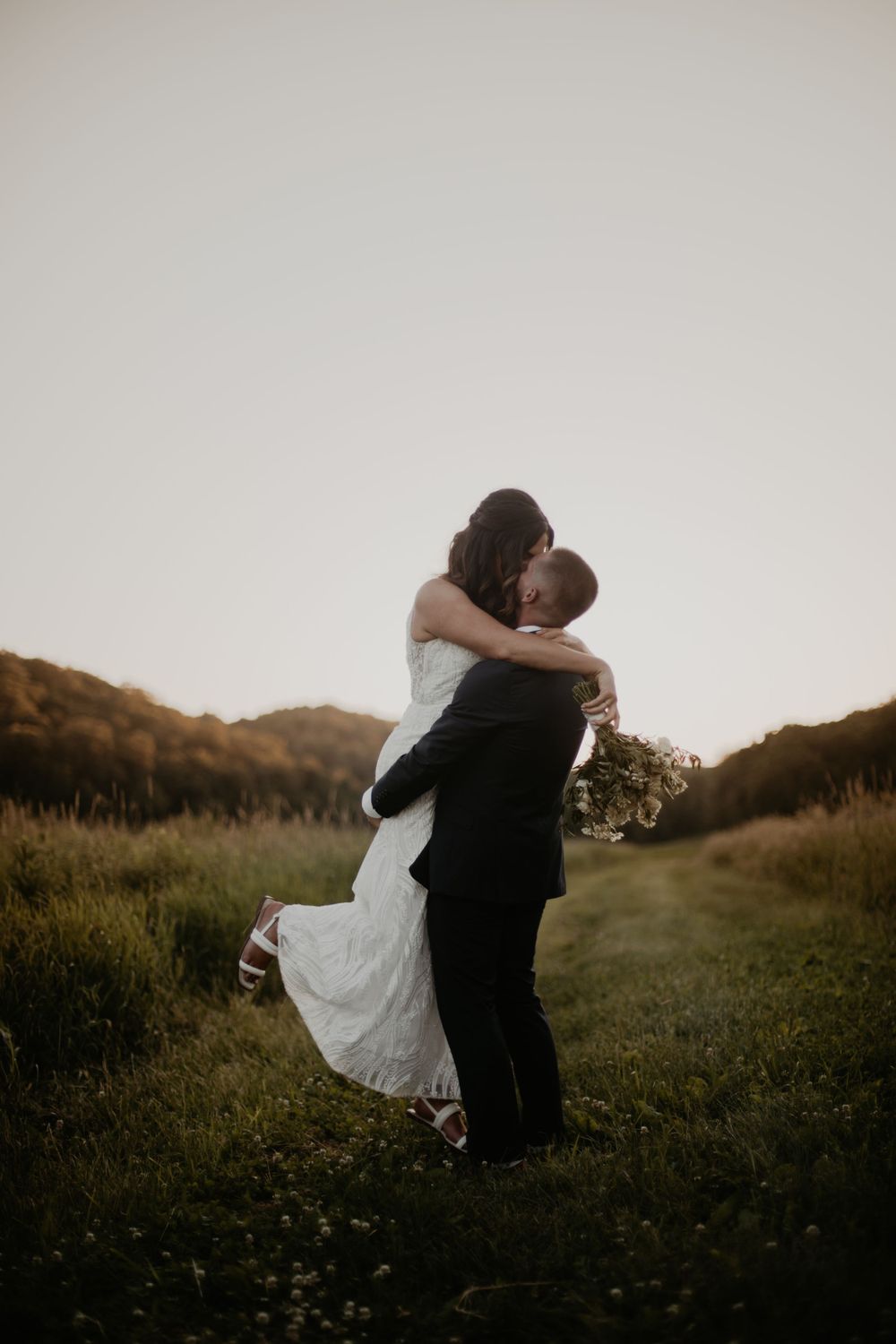 A couple embraces in a grassy field during golden hour with rolling hills in the background.