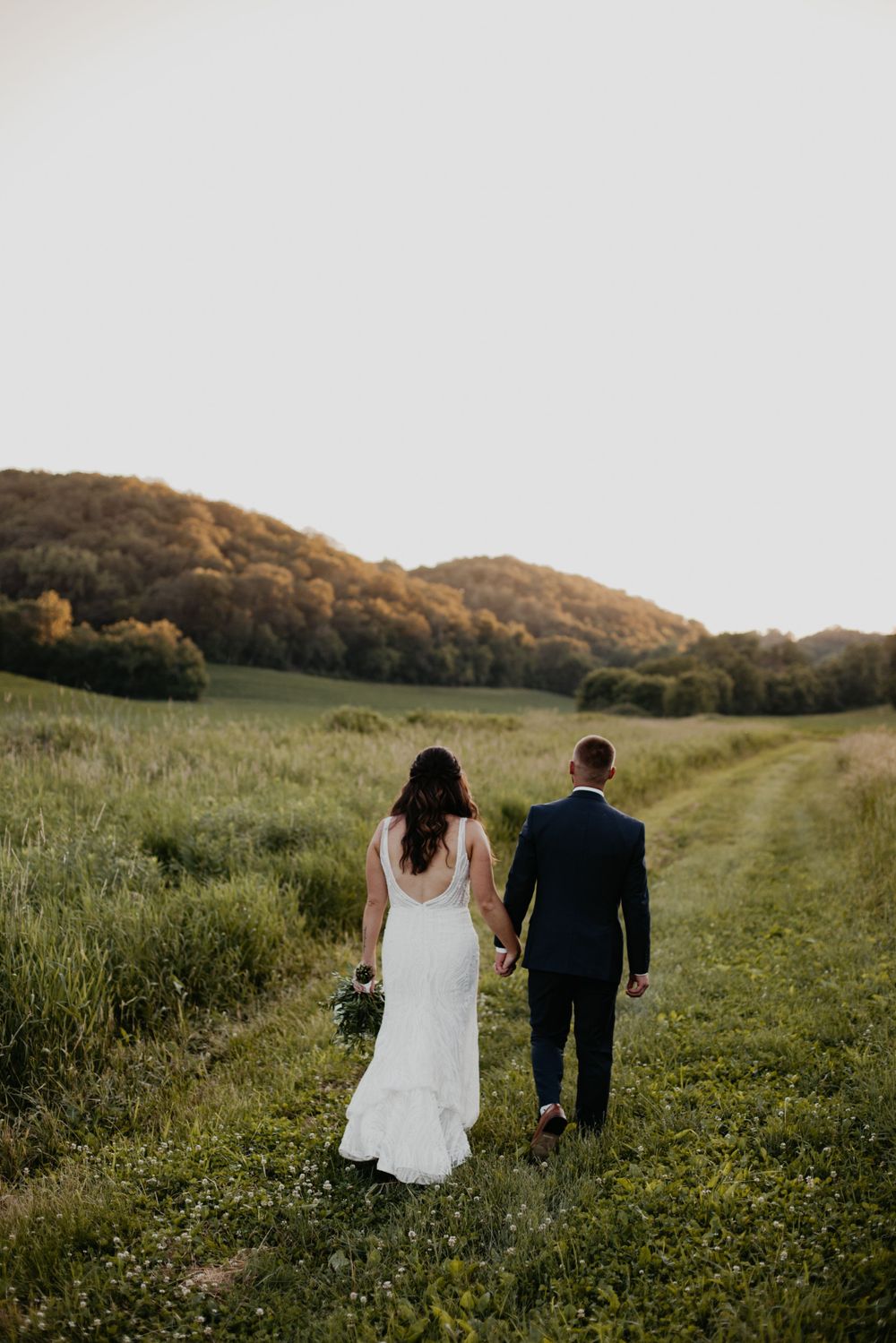 A couple in wedding attire walk hand in hand through a scenic green meadow with rolling hills in the background.