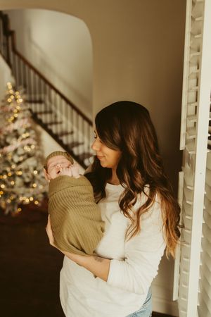 Brunette mother tenderly holding her swaddled newborn baby near a window with a Christmas tree.