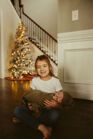 Young girl sitting cross-legged on hardwood floor holding a swaddled newborn in front of a Christmas tree.