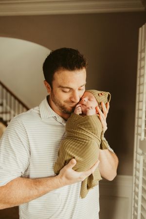 Father gently kissing his swaddled newborn baby wrapped in an olive green blanket indoors.