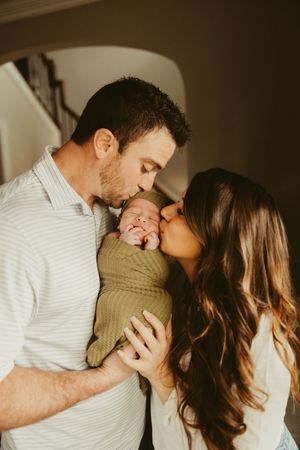 Parents kissing their smiling swaddled newborn baby together in a warmly lit home.