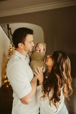 New parents share a tender moment holding their swaddled newborn in olive green wrap near Christmas tree lights.