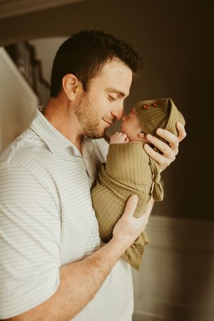 New father sharing a gentle nose-to-nose moment with his swaddled newborn in olive green wrap.