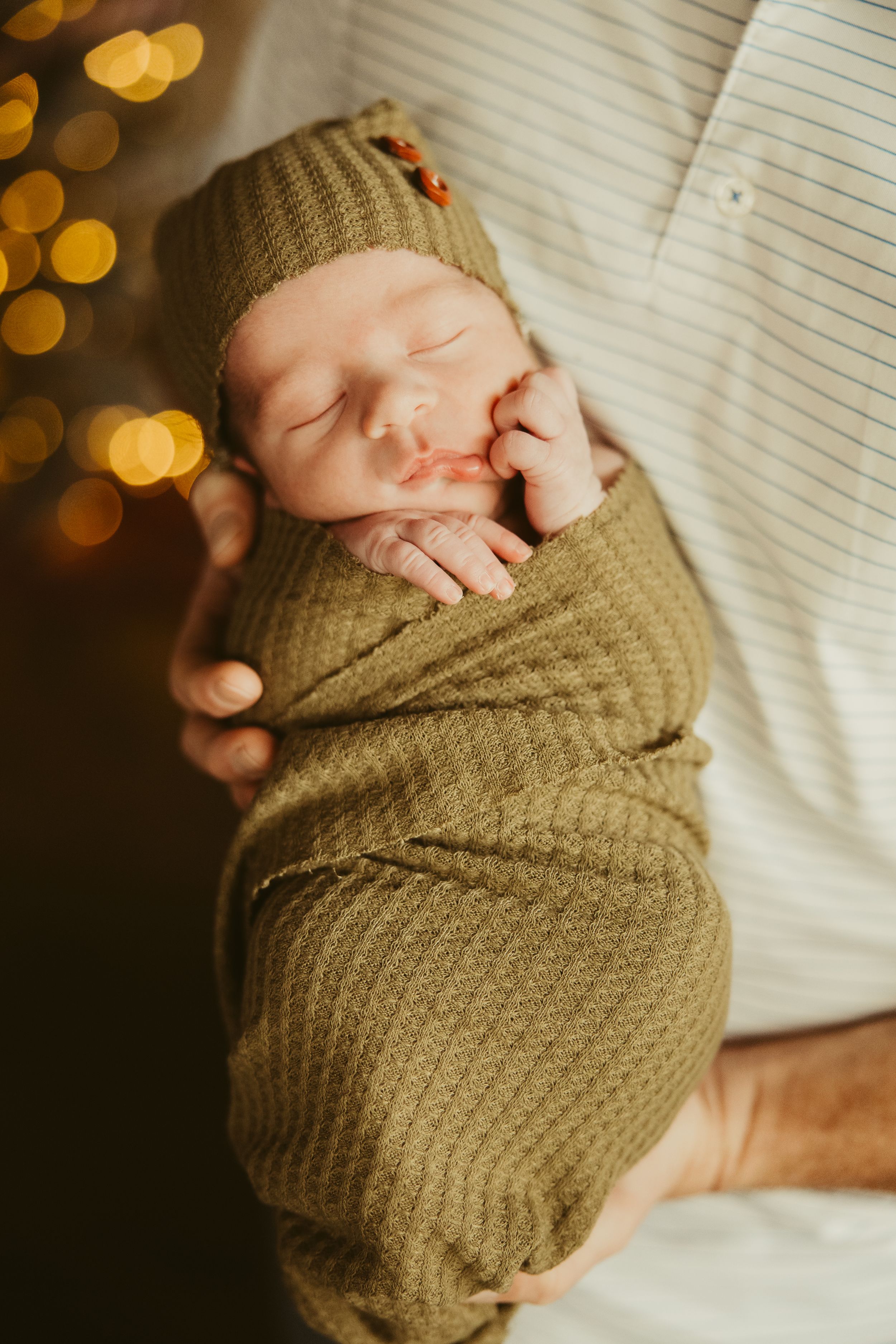 Sleeping newborn baby swaddled in olive green knit wrap with matching knotted hat held by father.