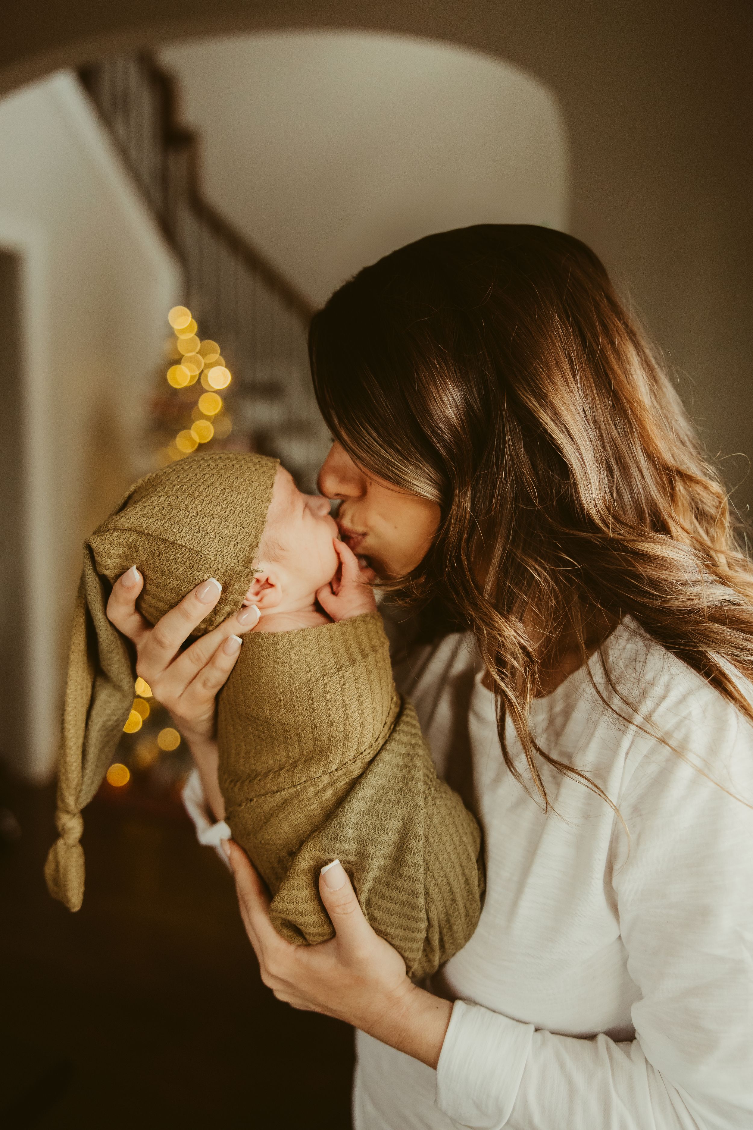 New mother giving a tender kiss to her swaddled newborn in olive green wrap with bokeh lights.