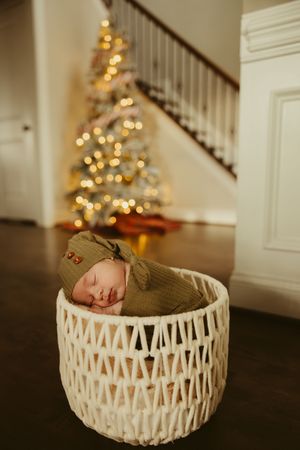 Sleeping newborn in olive green swaddle resting in white woven basket before lit Christmas tree.