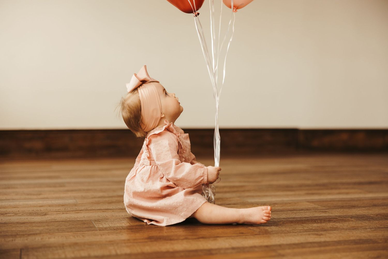 Adorable baby girl in pink dress sitting on wood floor holding rose gold balloons.