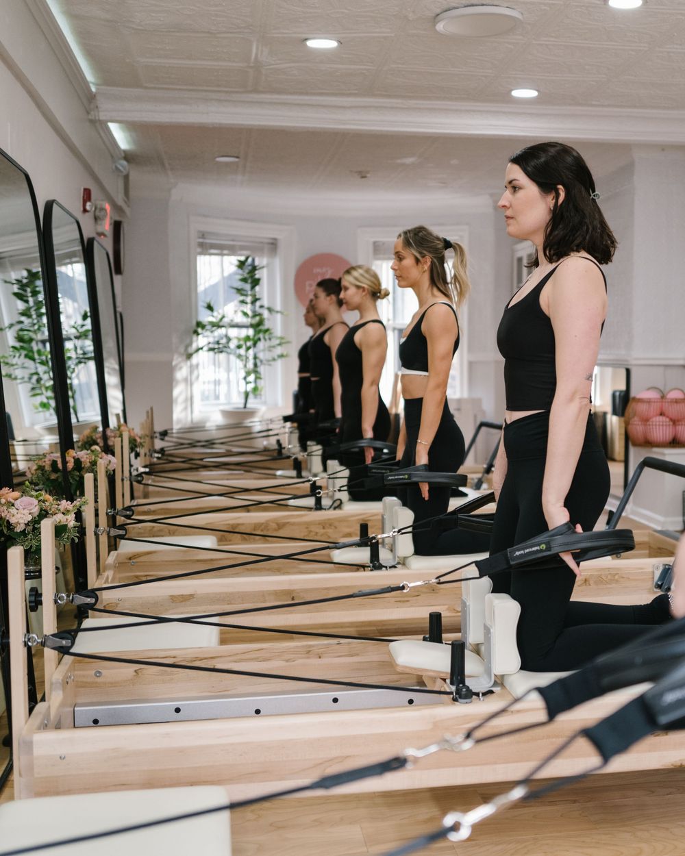 A group of people practicing Pilates exercises on reformer machines in a bright, modern studio with mirrored walls.