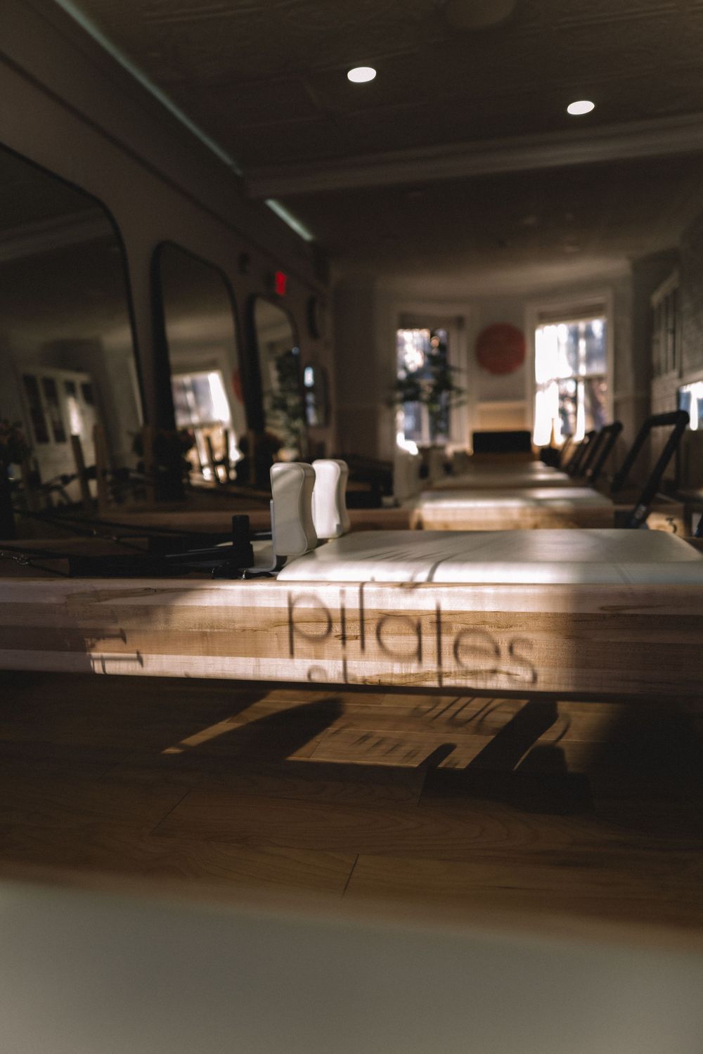 Pilates studio interior with sunlight streaming through windows, showing exercise equipment in a dimly lit space.