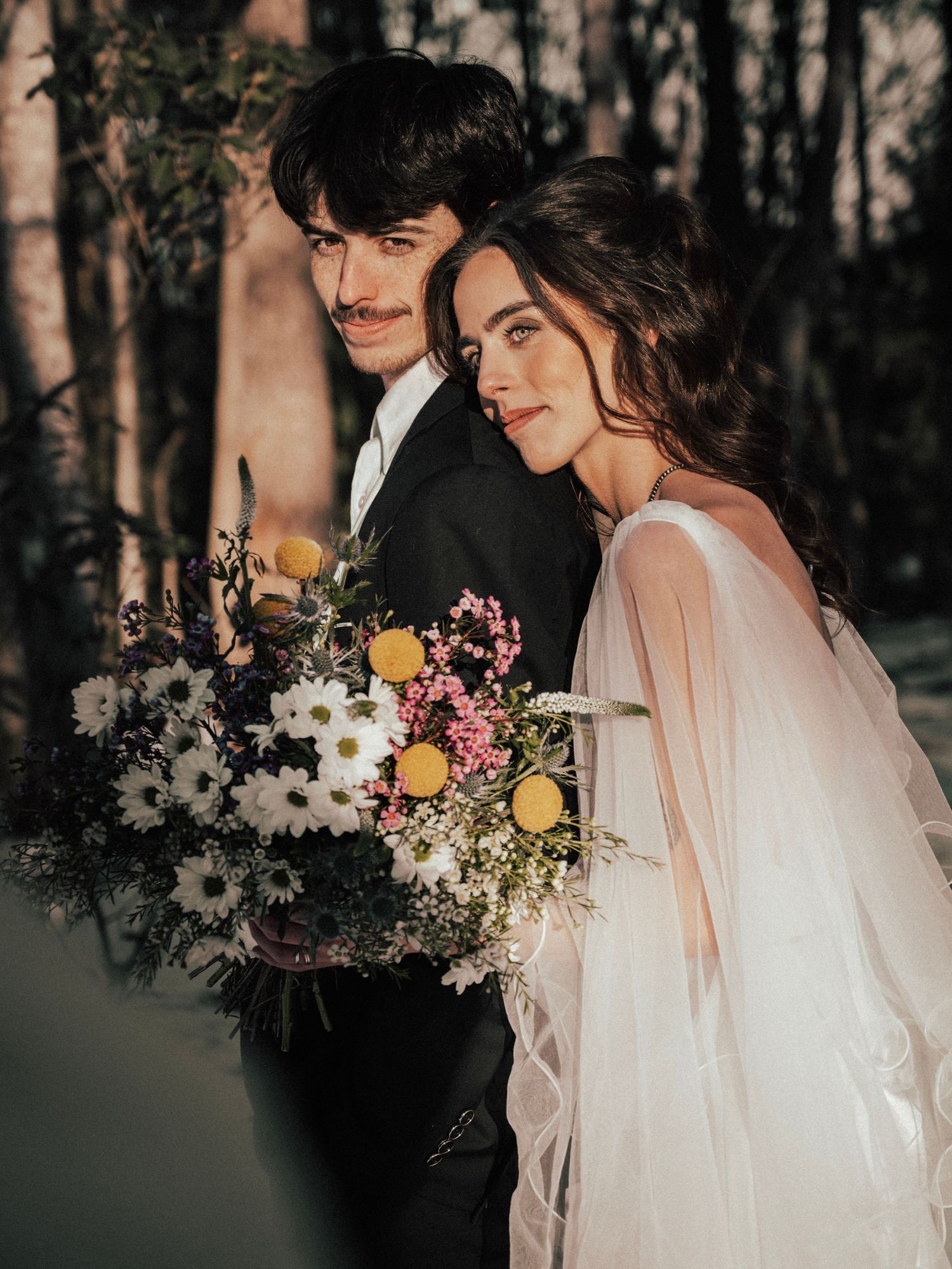 Ethereal wedding portrait with flowing veil and colorful wildflower bouquet in forest setting.