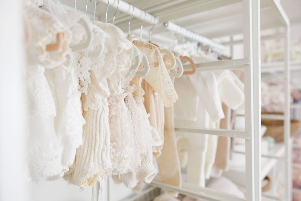Delicate white and cream baby clothes hanging neatly on a rack in a bright, organized nursery closet.