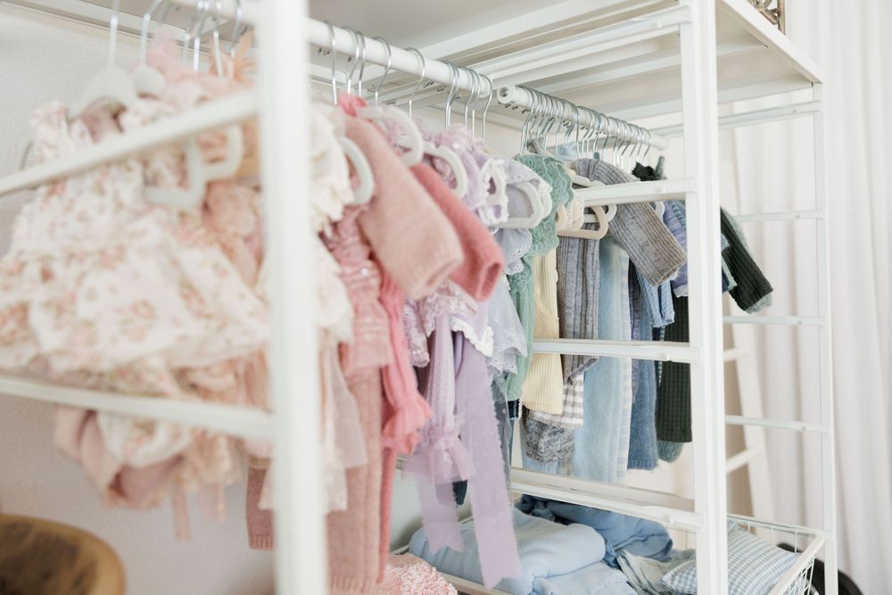 White rack displaying pastel baby clothes including pink floral dresses and blue garments in a bright nursery closet.
