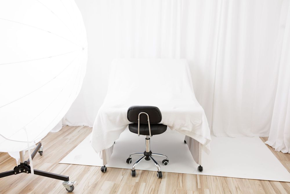 Professional photo studio setup with white backdrop, padded table, and black office chair on wood floor.