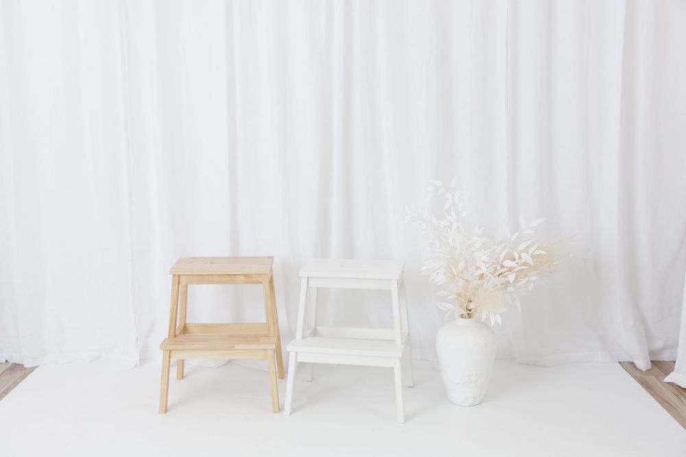 Two wooden step stools beside a white vase with dried flowers against a white curtain backdrop.