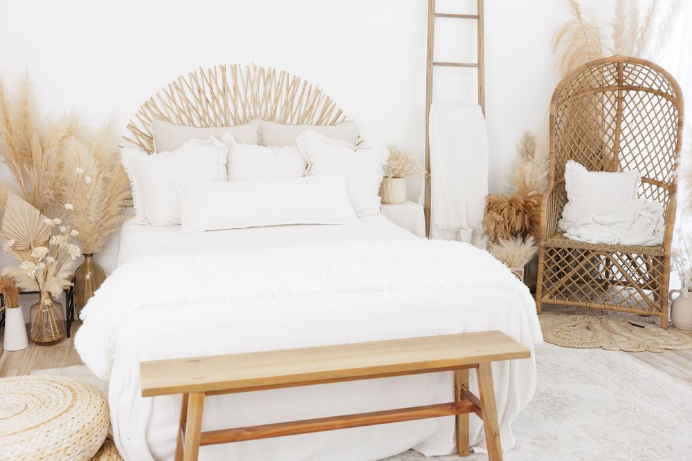 Boho bedroom with white bedding, rattan headboard, pampas grass, wicker chair, and wooden bench.