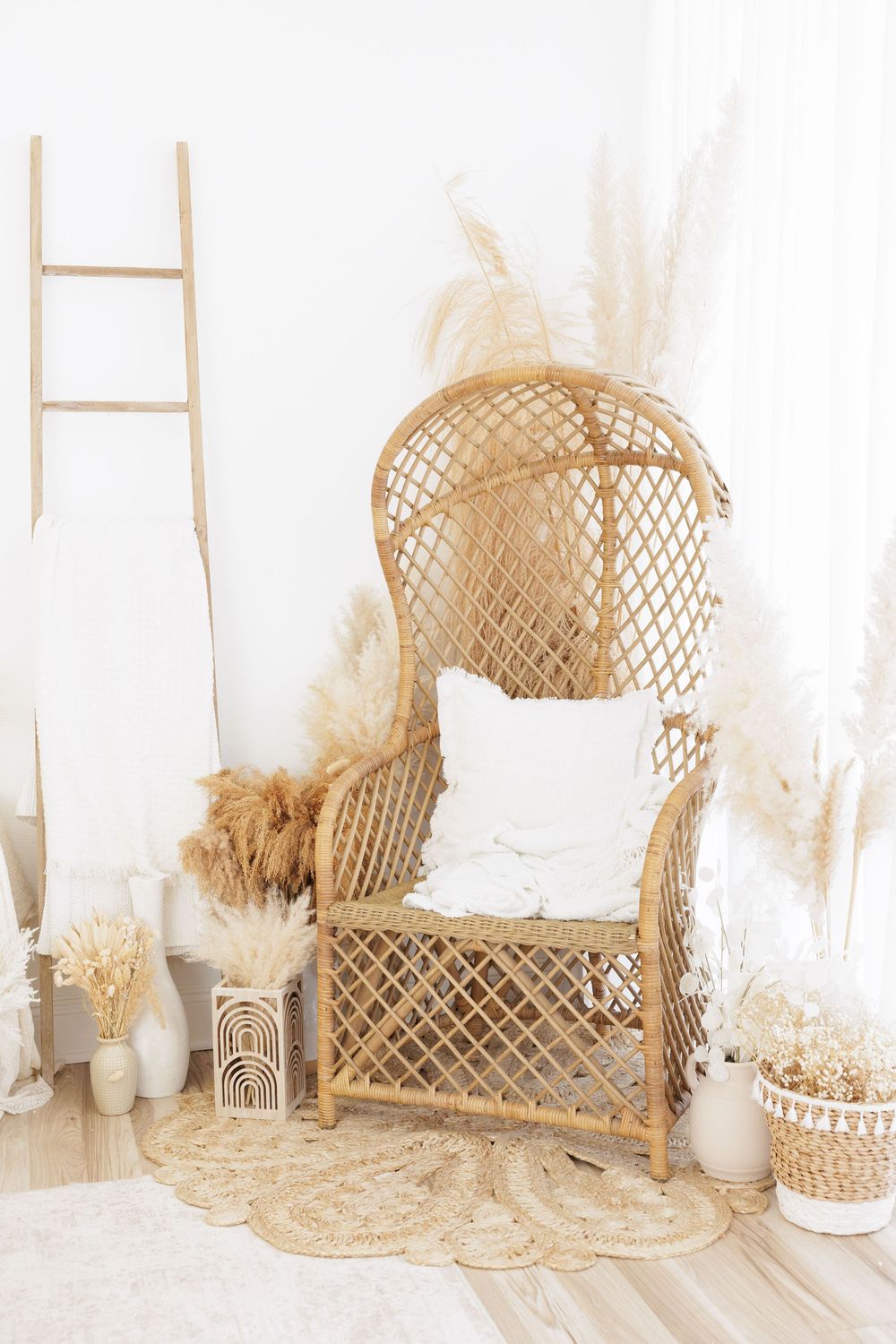 Boho-style rattan peacock chair with white pillow, pampas grass, dried botanicals, and a wooden ladder in a bright room.