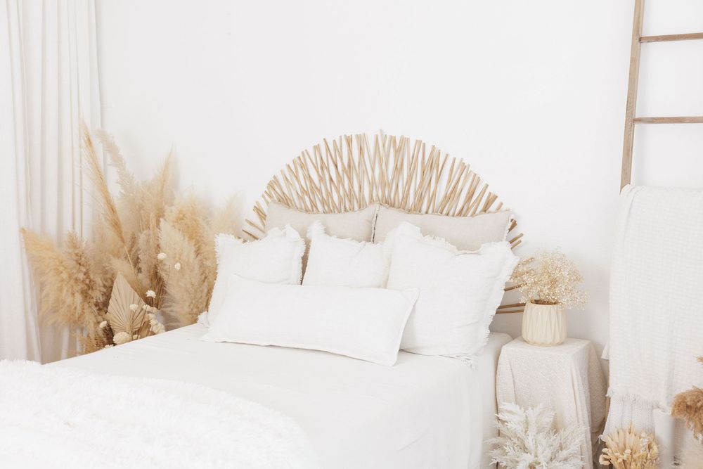 A serene boho-style white bed with decorative dried pampas grass and a woven rattan headboard in a bright room.