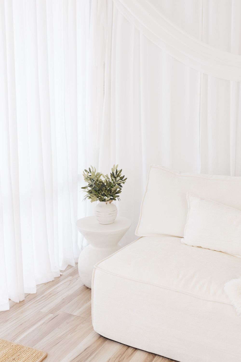 Minimalist white living room with a plush cream sofa, sheer curtains, and a small plant on a round side table.