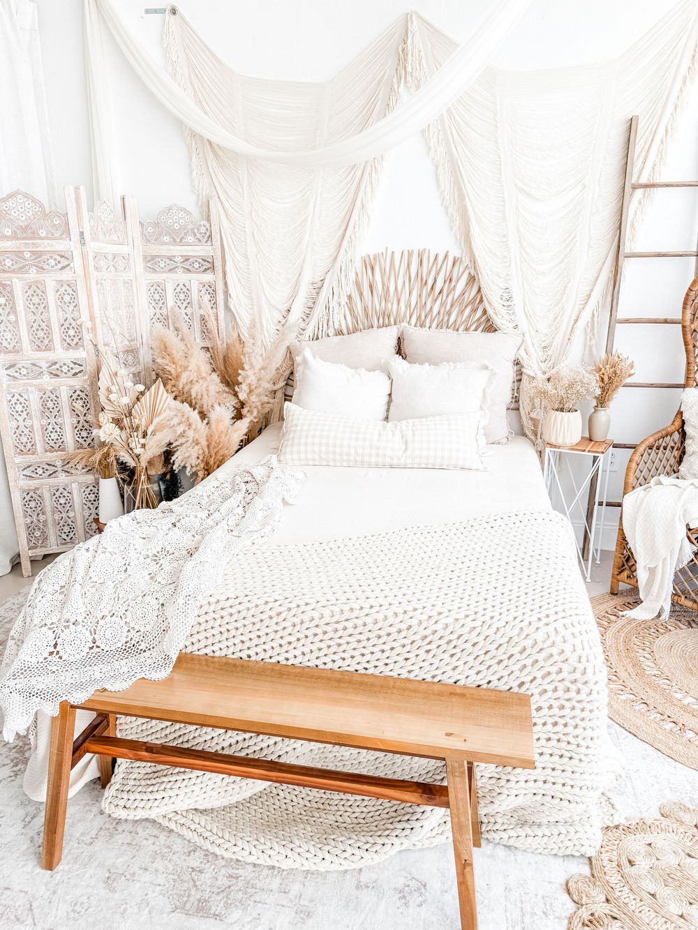 Boho bedroom with white bedding, macrame wall hanging, rattan peacock chair, and wooden bench in neutral tones.