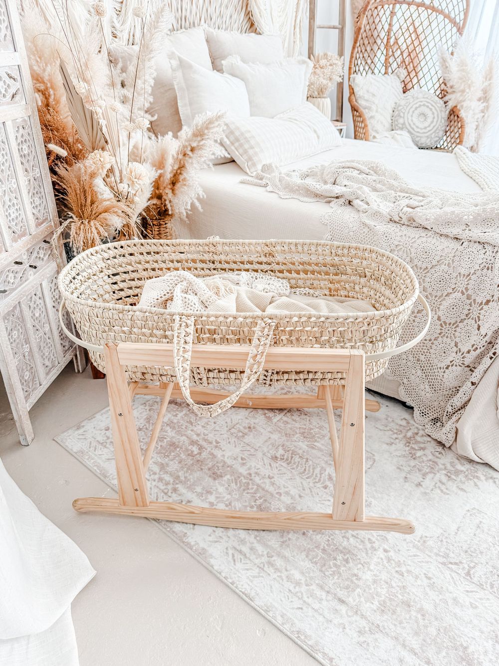 Boho-style woven bassinet with wooden rocking base surrounded by white bedding and pampas grass decor.