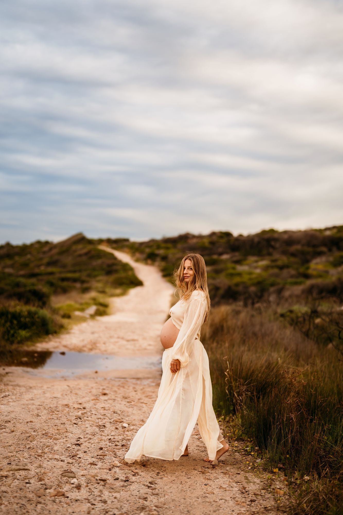 A white flowy dress moves gracefully along a winding coastal path through natural brush on an overcast day.
