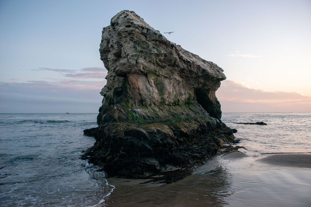 Panoramic sequence of a large rock formation on a beach at sunset with waves crashing against the shore.