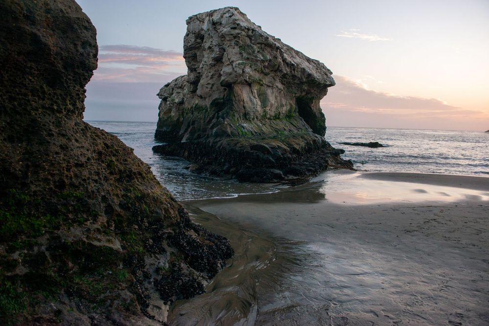 Large rock formations stand against a misty sunset at a rugged coastal beach with waves rolling onto shore.