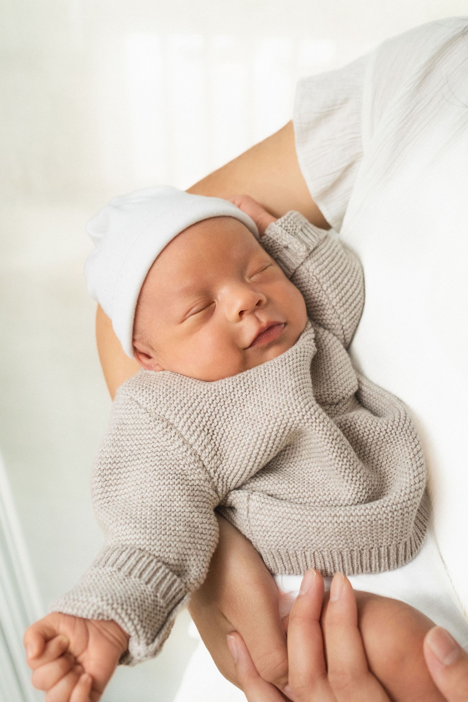 Sleeping newborn baby in beige knit outfit and white hat cradled in mother's arms.
