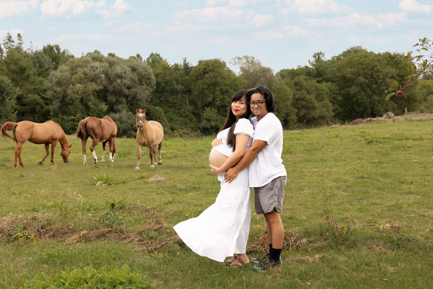 A couple poses together in a field with wild horses grazing in the background during a photo session.
